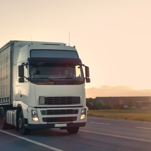 Front-View of Semi-Truck with Cargo Trailer Driving on a Highway. He's Speeding Through Industrial Warehouse Area with Sunset in the Background.