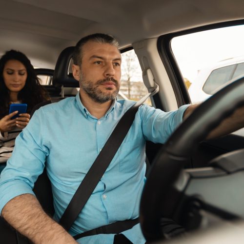 Portrait of focused male driver riding car, beautiful lady sitting inside auto on back passenger seat, using cell phone, having safe ride in the city, windshield view